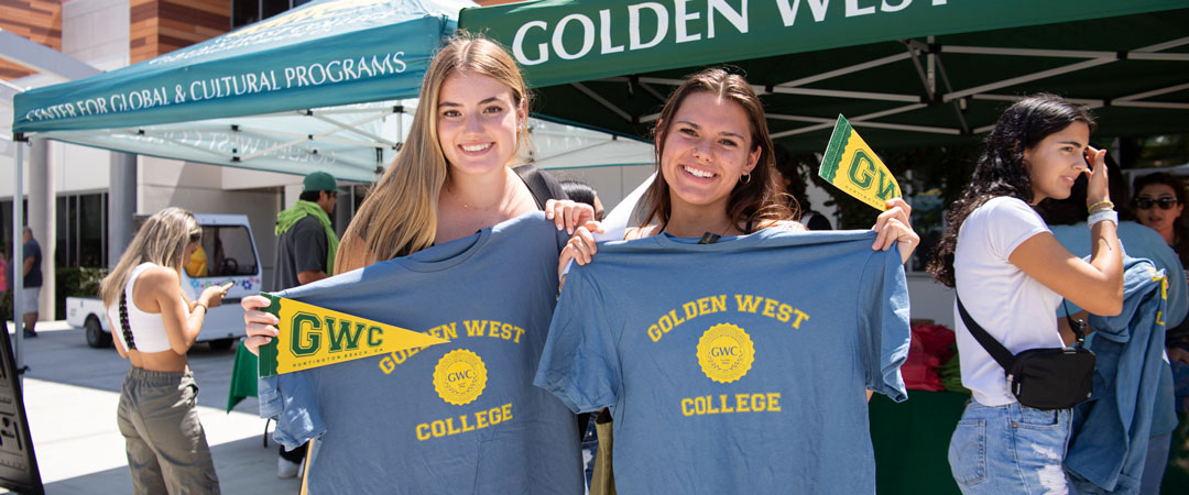 Two students hold up Golden West College shirts while standing in front of a promotional tent.