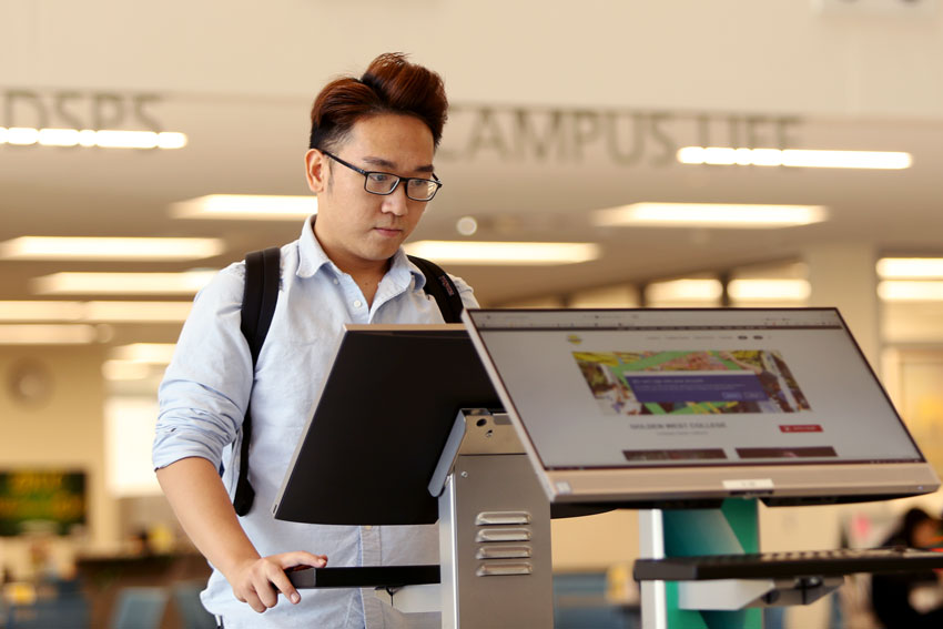 Student wearing glasses and a backpack uses a standing computer kiosk in a campus building.