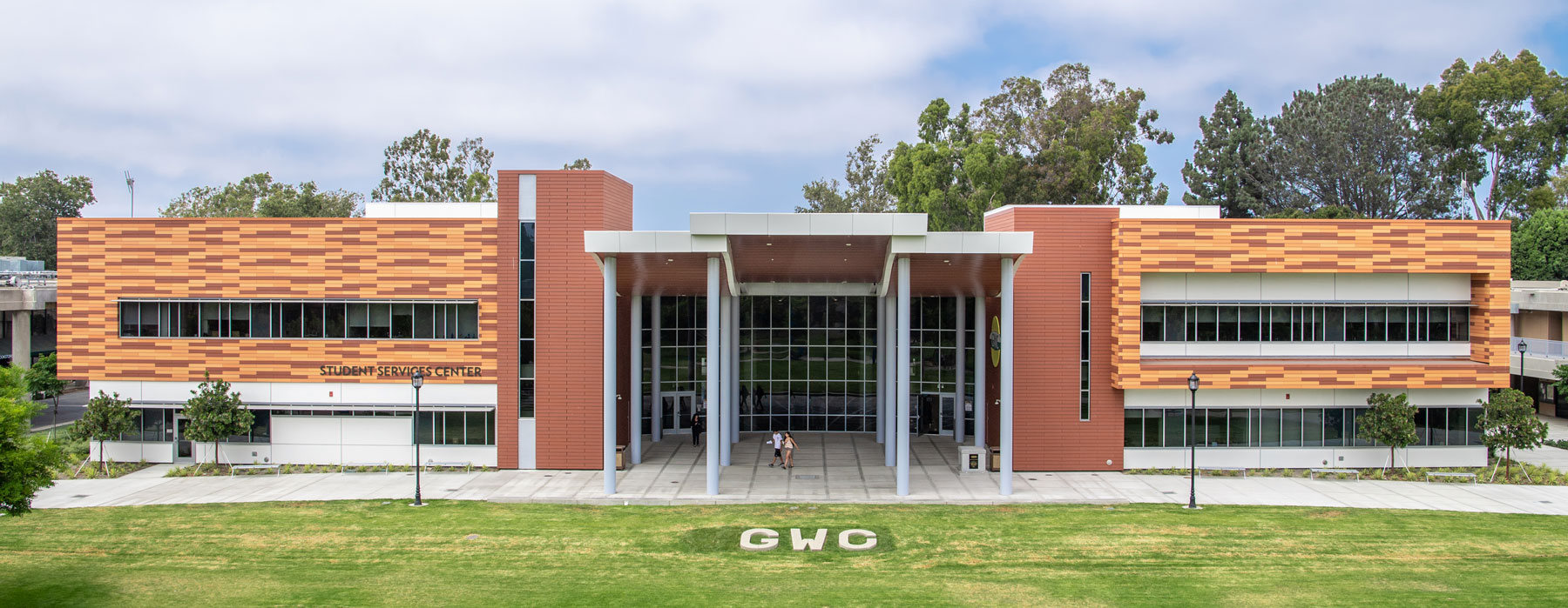 Modern campus building with a glass-fronted entrance and orange patterned facade labeled "Student Services Center," with a grassy lawn in front.