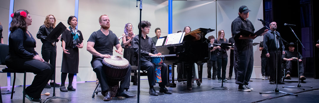 A choir performance featuring singers, musicians playing a drum and piano, and a speaker on stage, all dressed in black attire.